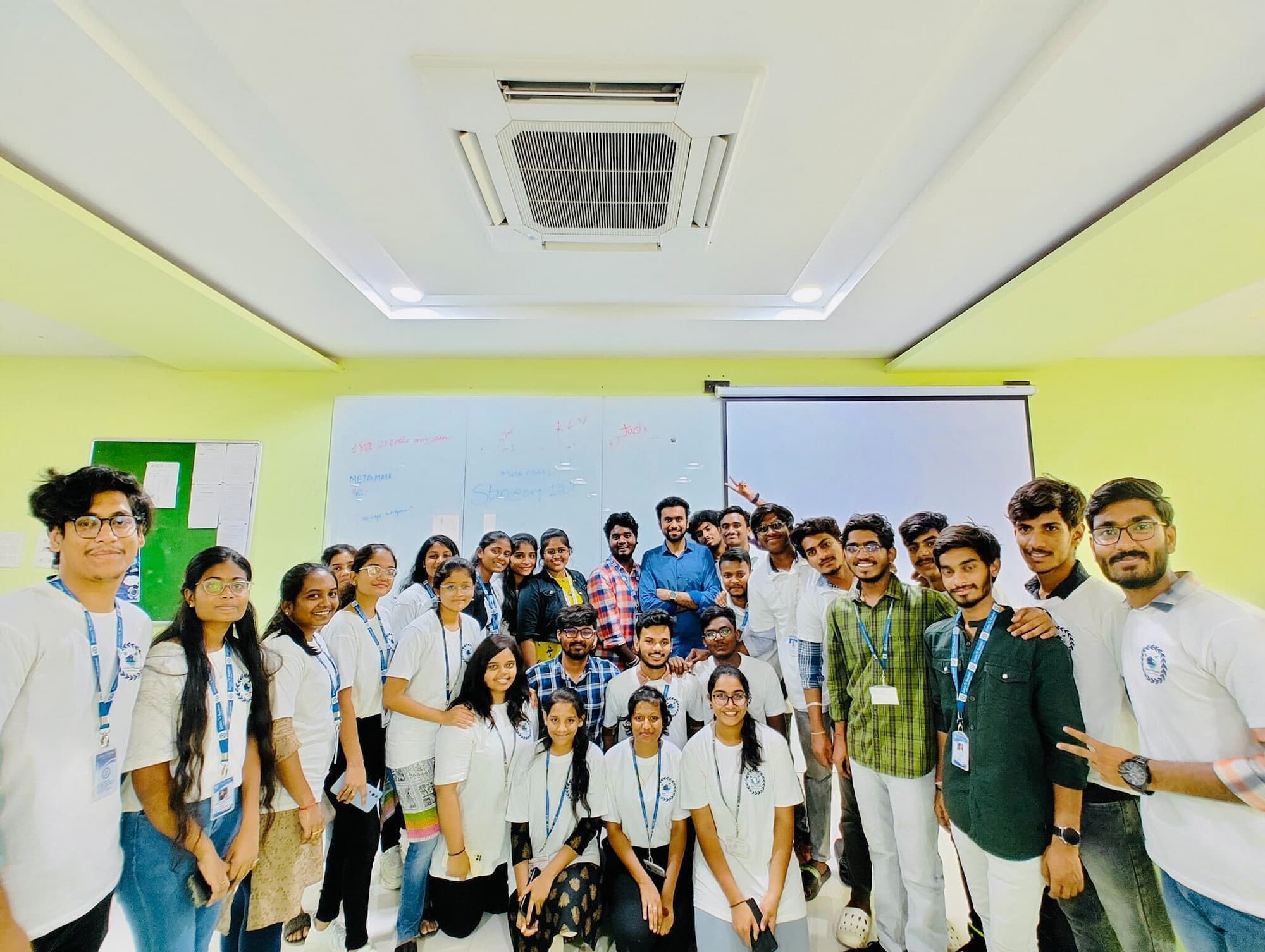 A large group of students and organizers posing for a group photo in a classroom in India. They are all smiling and wearing ID badges, with some in white shirts featuring a logo. The room has a bright yellow-green color with a whiteboard and projector screen in the background.